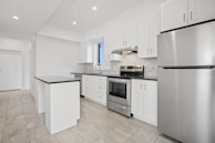 A modern kitchen featuring white cabinetry, stainless steel appliances, and a sleek black countertop. The room is well-lit with recessed lighting and has large floor tiles that add to the spacious feel. A small island provides additional workspace.