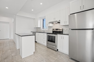 A modern kitchen featuring white cabinetry, stainless steel appliances, and a sleek black countertop. The room is well-lit with recessed lighting and has large floor tiles that add to the spacious feel. A small island provides additional workspace.