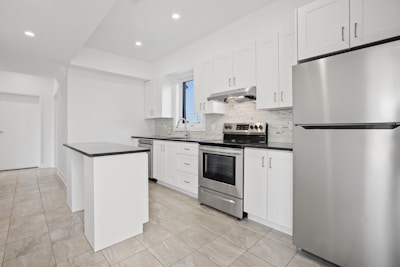 A modern kitchen featuring white cabinetry, stainless steel appliances, and a sleek black countertop. The room is well-lit with recessed lighting and has large floor tiles that add to the spacious feel. A small island provides additional workspace.
