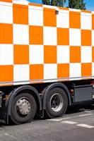 A large vehicle featuring a bright orange and white checkered pattern is parked on a street. The focus is on the side of the truck and its wheels. The ground is marked with road lines, and there are trees visible in the background.