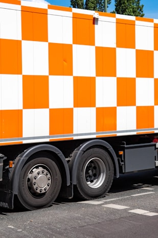 A large vehicle featuring a bright orange and white checkered pattern is parked on a street. The focus is on the side of the truck and its wheels. The ground is marked with road lines, and there are trees visible in the background.