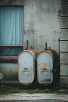 Two old, rusted metal tanks stand against a worn concrete wall next to a window with a blue curtain. The tanks are weathered and rusty, with grass growing at their base.