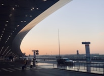 A modern transportation hub during sunset. The architecture features a sweeping curved roof with lights, a smooth roadway, and travelers with luggage crossing the street. A few cars are parked along the side, while directional signs are visible.