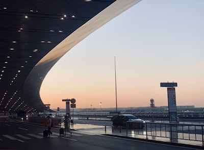 A modern transportation hub during sunset. The architecture features a sweeping curved roof with lights, a smooth roadway, and travelers with luggage crossing the street. A few cars are parked along the side, while directional signs are visible.