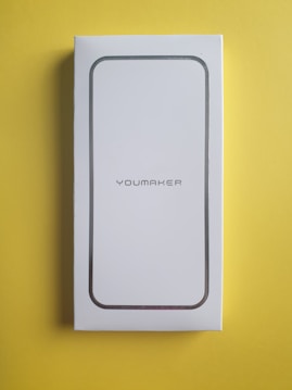 A white rectangular box, possibly packaging for a product, rests on a bright yellow surface. The box has rounded corners with a metallic outline and the word 'YOUMAKER' printed in the center.