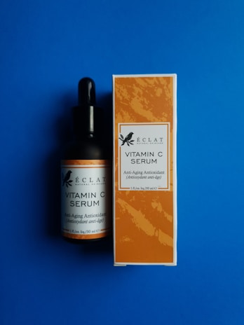 A bottle and a box of Éclat Natural Skincare Vitamin C Serum are placed against a solid blue background. The bottle has a black dropper cap, and the label matches the design on the box, which features an orange pattern with text indicating it is an anti-aging antioxidant product.