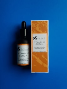 A bottle and a box of &Eacute;clat Natural Skincare Vitamin C Serum are placed against a solid blue background. The bottle has a black dropper cap, and the label matches the design on the box, which features an orange pattern with text indicating it is an anti-aging antioxidant product.