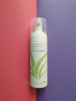 A bottle of aloe vera facial cleansing foam placed vertically against a split background of pink and purple. The bottle has a transparent cap and green aloe vera leaf graphics on its white surface.