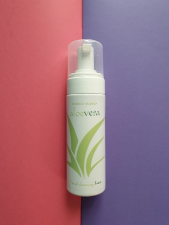 A bottle of aloe vera facial cleansing foam placed vertically against a split background of pink and purple. The bottle has a transparent cap and green aloe vera leaf graphics on its white surface.