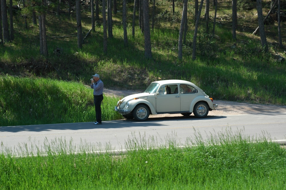 man in black shirt and blue denim jeans standing beside white sedan on road during daytime