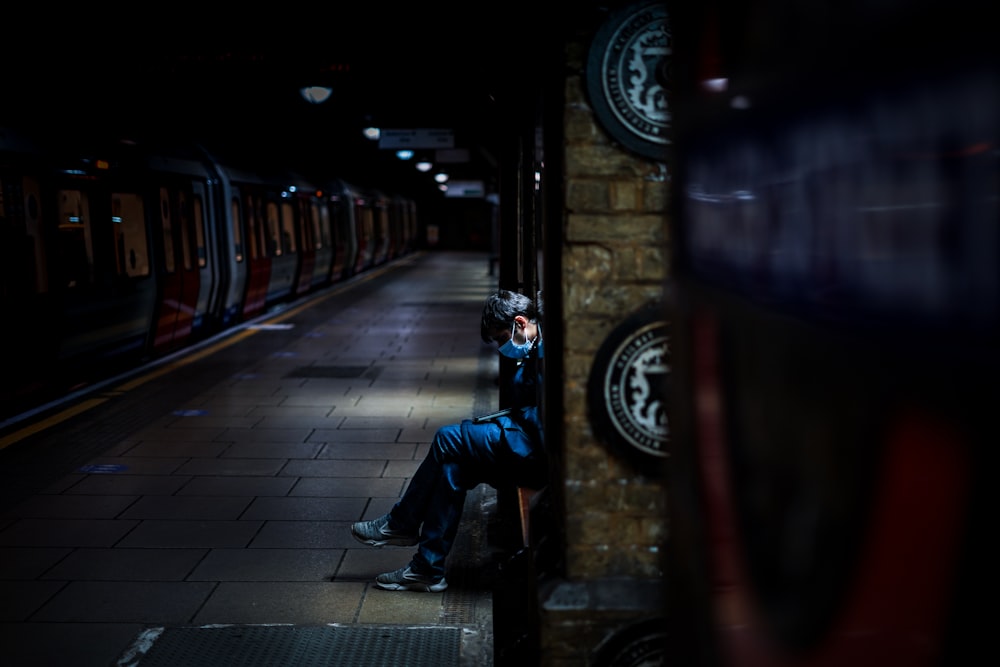 man in black jacket and blue denim jeans sitting on sidewalk during night time