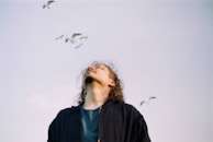 A person with long hair is gazing upwards with a peaceful expression, surrounded by birds flying in a clear sky. The person is wearing a dark jacket and a blue shirt, and appears thoughtful and serene.