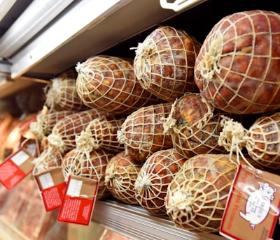 A display of cured meats wrapped in netting and stacked neatly on a shelf, with some price tags attached.