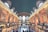 A large, bustling train station interior featuring a grand arched ceiling with celestial designs, tall arched windows allowing natural light to flood the space, and numerous people walking and gathering. An American flag hangs prominently on the right, and information boards and ticket counters line the walls.