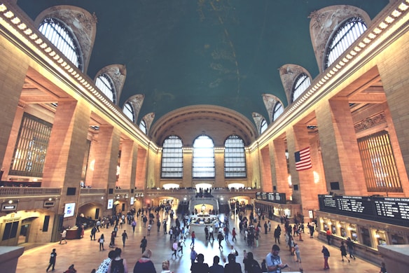 A large, bustling train station interior featuring a grand arched ceiling with celestial designs, tall arched windows allowing natural light to flood the space, and numerous people walking and gathering. An American flag hangs prominently on the right, and information boards and ticket counters line the walls.
