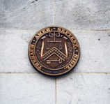 A bronze seal mounted on a light gray stone wall, featuring a shield with a set of scales, a chevron and chessboard pattern, and a key. The seal is inscribed with Latin text around its perimeter.