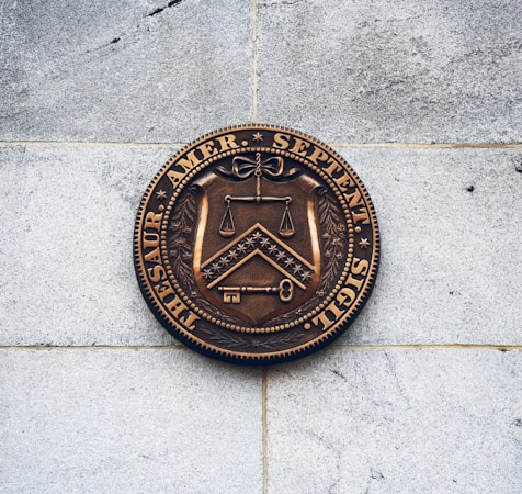 A bronze seal mounted on a light gray stone wall, featuring a shield with a set of scales, a chevron and chessboard pattern, and a key. The seal is inscribed with Latin text around its perimeter.
