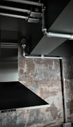 The image depicts an industrial-style interior space with exposed metal piping on a ceiling. There is a stainless steel vent hood and a textured wall with a rustic, weathered appearance.