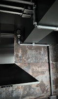 The image depicts an industrial-style interior space with exposed metal piping on a ceiling. There is a stainless steel vent hood and a textured wall with a rustic, weathered appearance.