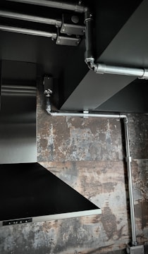 The image depicts an industrial-style interior space with exposed metal piping on a ceiling. There is a stainless steel vent hood and a textured wall with a rustic, weathered appearance.