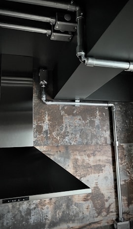 The image depicts an industrial-style interior space with exposed metal piping on a ceiling. There is a stainless steel vent hood and a textured wall with a rustic, weathered appearance.