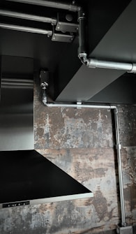 The image depicts an industrial-style interior space with exposed metal piping on a ceiling. There is a stainless steel vent hood and a textured wall with a rustic, weathered appearance.