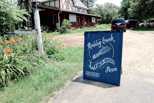 A blue signboard with white lettering stands in front of a rustic wooden house. The sign reads 'Rowley Creek Lavender Farm' and includes an illustration of lavender. The house has a porch and is surrounded by greenery, with a few orange flowers nearby. There are cars parked on a dirt driveway beside the house.
