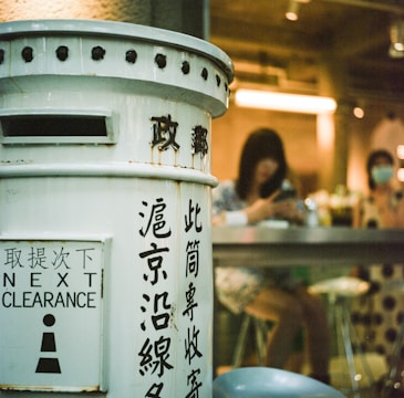 A vintage-style mailbox painted white with black text stands prominently in the foreground. The text includes various characters and 'Next Clearance' written in English. In the background, two people are seated at a high counter inside a dimly lit space, with one person looking at a phone. The interior features warm lighting.