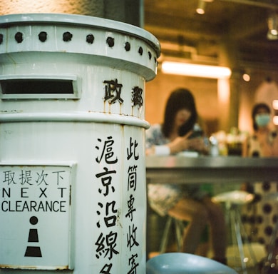 A vintage-style mailbox painted white with black text stands prominently in the foreground. The text includes various characters and 'Next Clearance' written in English. In the background, two people are seated at a high counter inside a dimly lit space, with one person looking at a phone. The interior features warm lighting.