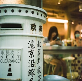 A vintage-style mailbox painted white with black text stands prominently in the foreground. The text includes various characters and 'Next Clearance' written in English. In the background, two people are seated at a high counter inside a dimly lit space, with one person looking at a phone. The interior features warm lighting.