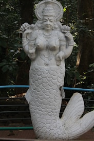 A white stone statue of a mermaid-like goddess, adorned with intricate jewelry and a headdress, set against a lush green forest background. The statue stands upright with one hand raised in a gesture of blessing.