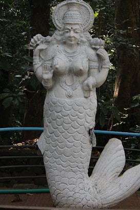 A white stone statue of a mermaid-like goddess, adorned with intricate jewelry and a headdress, set against a lush green forest background. The statue stands upright with one hand raised in a gesture of blessing.