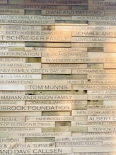 A wall displaying various wooden plaques with engraved names and messages, likely representing donors or memorials. The plaques are arranged in a brick-like pattern, with each one varying slightly in wood tone and text size.