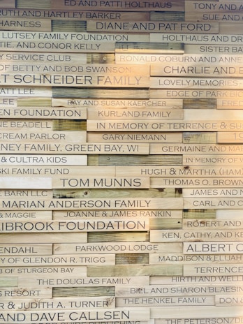 A wall displaying various wooden plaques with engraved names and messages, likely representing donors or memorials. The plaques are arranged in a brick-like pattern, with each one varying slightly in wood tone and text size.