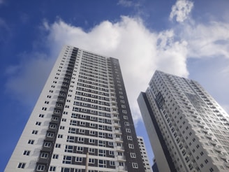 Two modern high-rise apartment buildings extend into the sky, with a clear blue sky and scattered clouds in the background. The buildings have a mix of dark and light-colored panels and numerous windows.