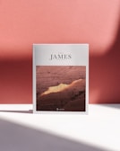 A book titled 'Book of James' stands upright against a soft pink background. The cover features an image of a desert landscape with rugged, earthy textures and warm lighting. The shadows on the surface suggest bright, directional light. The book's positioning and shadows add a sense of depth and dimension.