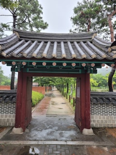 A traditional wooden gate with ornate roof tiles leads to a long pathway surrounded by trees and greenery. The gate is detailed with colorful floral motifs under the eaves. Brick and stone walls line the path, and tall trees flank the walkway, creating a serene and natural ambiance.