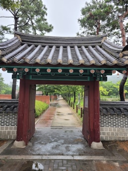 A traditional wooden gate with ornate roof tiles leads to a long pathway surrounded by trees and greenery. The gate is detailed with colorful floral motifs under the eaves. Brick and stone walls line the path, and tall trees flank the walkway, creating a serene and natural ambiance.