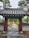 A traditional wooden gate with ornate roof tiles leads to a long pathway surrounded by trees and greenery. The gate is detailed with colorful floral motifs under the eaves. Brick and stone walls line the path, and tall trees flank the walkway, creating a serene and natural ambiance.
