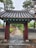 A traditional wooden gate with ornate roof tiles leads to a long pathway surrounded by trees and greenery. The gate is detailed with colorful floral motifs under the eaves. Brick and stone walls line the path, and tall trees flank the walkway, creating a serene and natural ambiance.