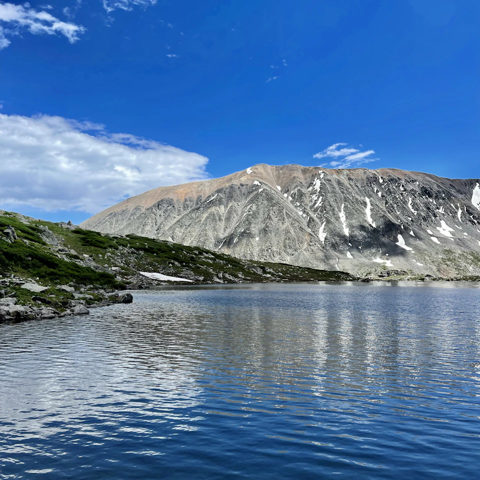 The image depicts a serene mountain lake nestled amidst a lush, green landscape. The calm waters of the lake reflect the surrounding mountains and sky, creating a mirror-like effect that enhances the beauty of the scene. In the distance, snow-capped peaks rise majestically against the clear blue sky, adding depth to the image.

The perspective of the photo is from the shoreline, looking out towards the mountain range. The mountains in the background are covered in a blanket of snow, their white peaks contrasting with the blue sky and green vegetation.