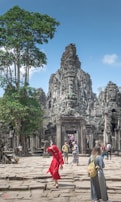 A group of tourists explores an ancient stone temple with intricate carvings, surrounded by lush greenery. One person in a vibrant red outfit stands out among the muted stone colors. The sky is clear and blue, adding a sense of openness to the scene.
