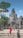 A group of tourists explores an ancient stone temple with intricate carvings, surrounded by lush greenery. One person in a vibrant red outfit stands out among the muted stone colors. The sky is clear and blue, adding a sense of openness to the scene.