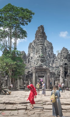 A group of tourists explores an ancient stone temple with intricate carvings, surrounded by lush greenery. One person in a vibrant red outfit stands out among the muted stone colors. The sky is clear and blue, adding a sense of openness to the scene.