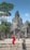A group of tourists explores an ancient stone temple with intricate carvings, surrounded by lush greenery. One person in a vibrant red outfit stands out among the muted stone colors. The sky is clear and blue, adding a sense of openness to the scene.