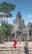 A group of tourists explores an ancient stone temple with intricate carvings, surrounded by lush greenery. One person in a vibrant red outfit stands out among the muted stone colors. The sky is clear and blue, adding a sense of openness to the scene.