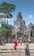 A group of tourists explores an ancient stone temple with intricate carvings, surrounded by lush greenery. One person in a vibrant red outfit stands out among the muted stone colors. The sky is clear and blue, adding a sense of openness to the scene.