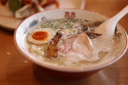 A bowl of ramen with rich broth, topped with a soft-boiled egg, slices of pork, and green onions. The bowl is detailed with traditional designs and a white soup spoon rests inside. The background shows a blurred dish on a wooden table.