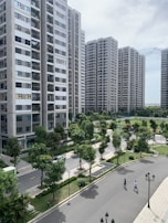 A series of modern high-rise apartment buildings surrounded by well-maintained green spaces and trees. The foreground features a winding road with two people walking, along with classic-style street lamps. The sky is partly cloudy, providing a bright yet calm ambiance.