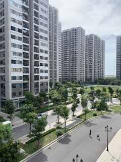 A series of modern high-rise apartment buildings surrounded by well-maintained green spaces and trees. The foreground features a winding road with two people walking, along with classic-style street lamps. The sky is partly cloudy, providing a bright yet calm ambiance.